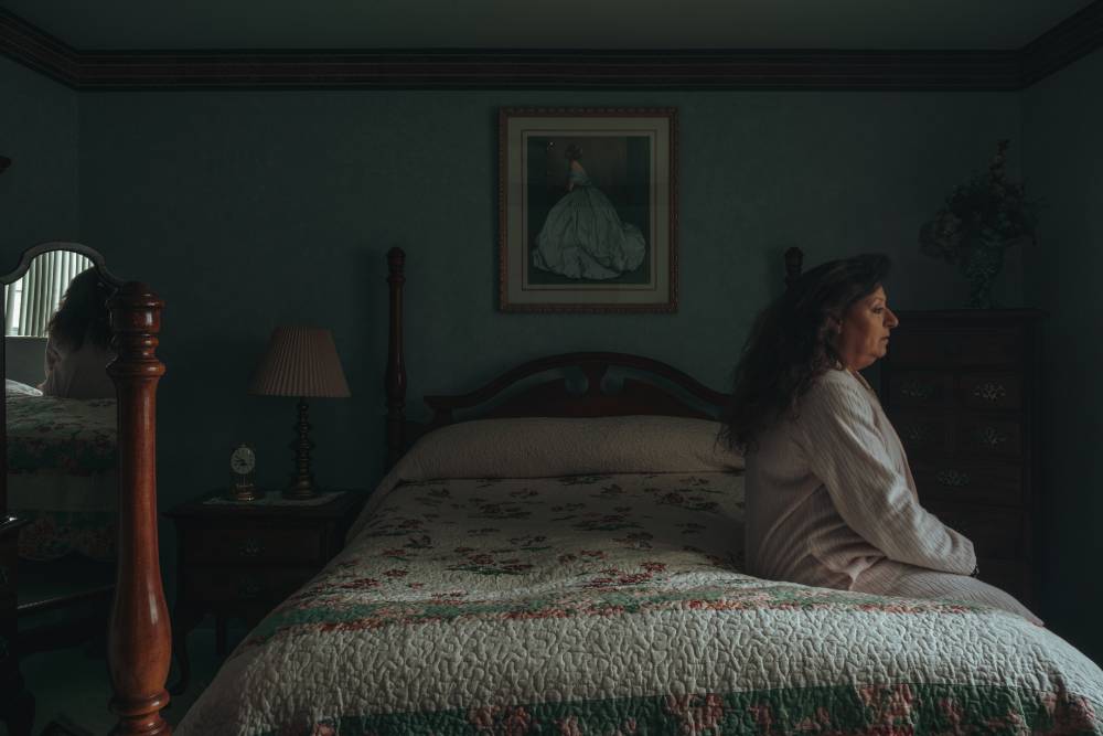 Side view of woman in bathrobe sitting on edge of bed in darkened room